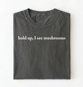 Flat lay of a pepper-colored t-shirt on a white background featuring brown text that reads “hold up, I see mushrooms. 