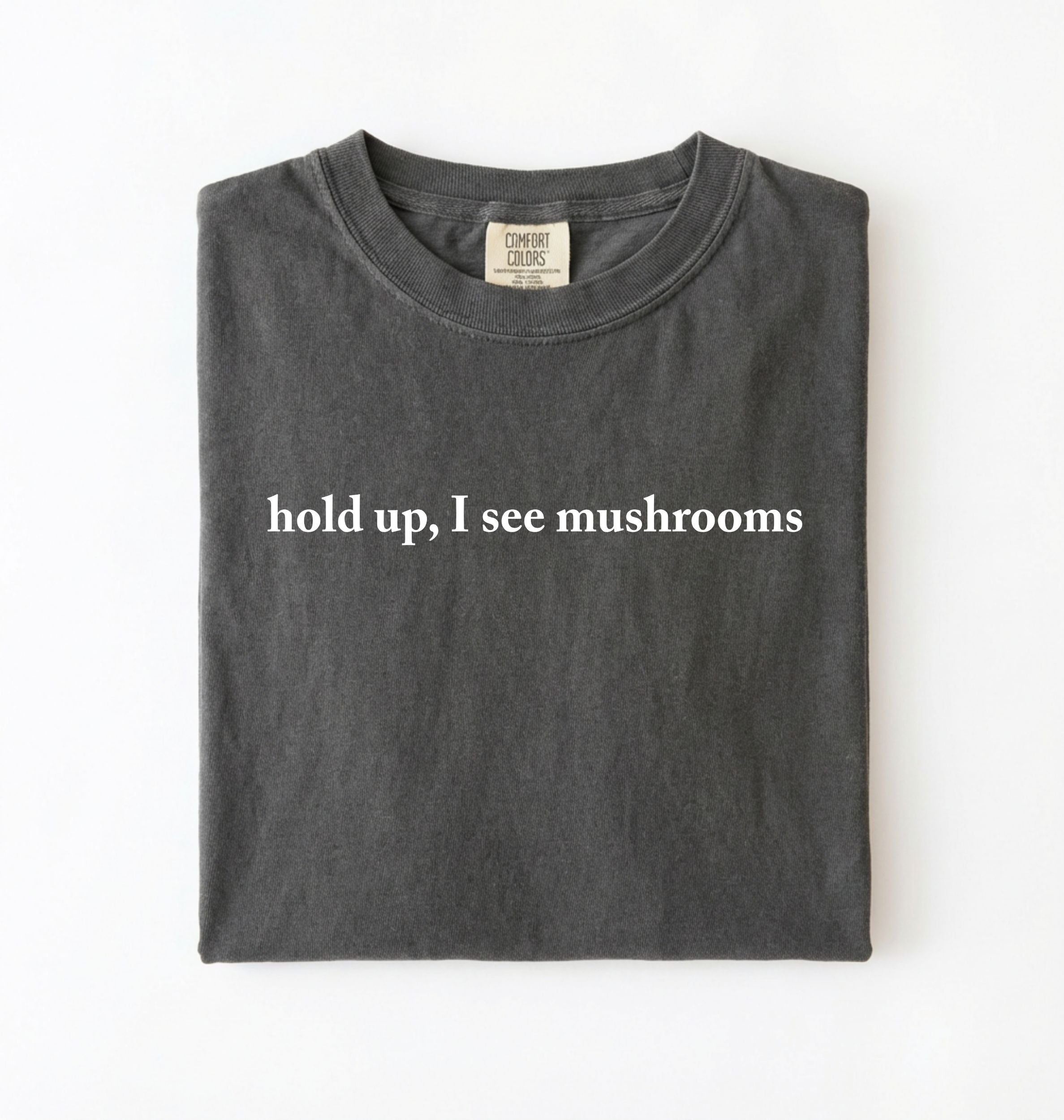 Flat lay of a pepper-colored t-shirt on a white background featuring brown text that reads “hold up, I see mushrooms. 