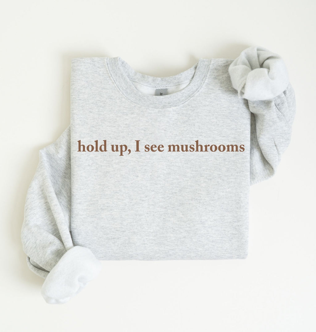 Flat lay of a ash-colored sweatshirt on a white background featuring brown text that reads “hold up, I see mushrooms. 