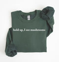 Flat lay of a forestgreen-colored sweatshirt on a white background featuring brown text that reads “hold up, I see mushrooms. 