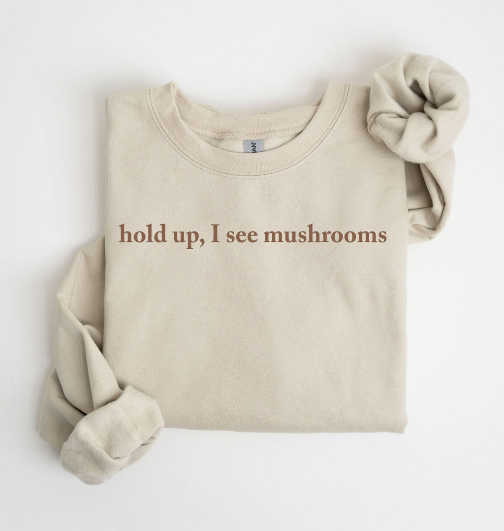 Flat lay of a sand-colored sweatshirt on a white background featuring brown text that reads “hold up, I see mushrooms. 