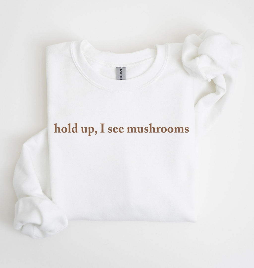 Flat lay of a white-colored sweatshirt on a white background featuring brown text that reads “hold up, I see mushrooms. 