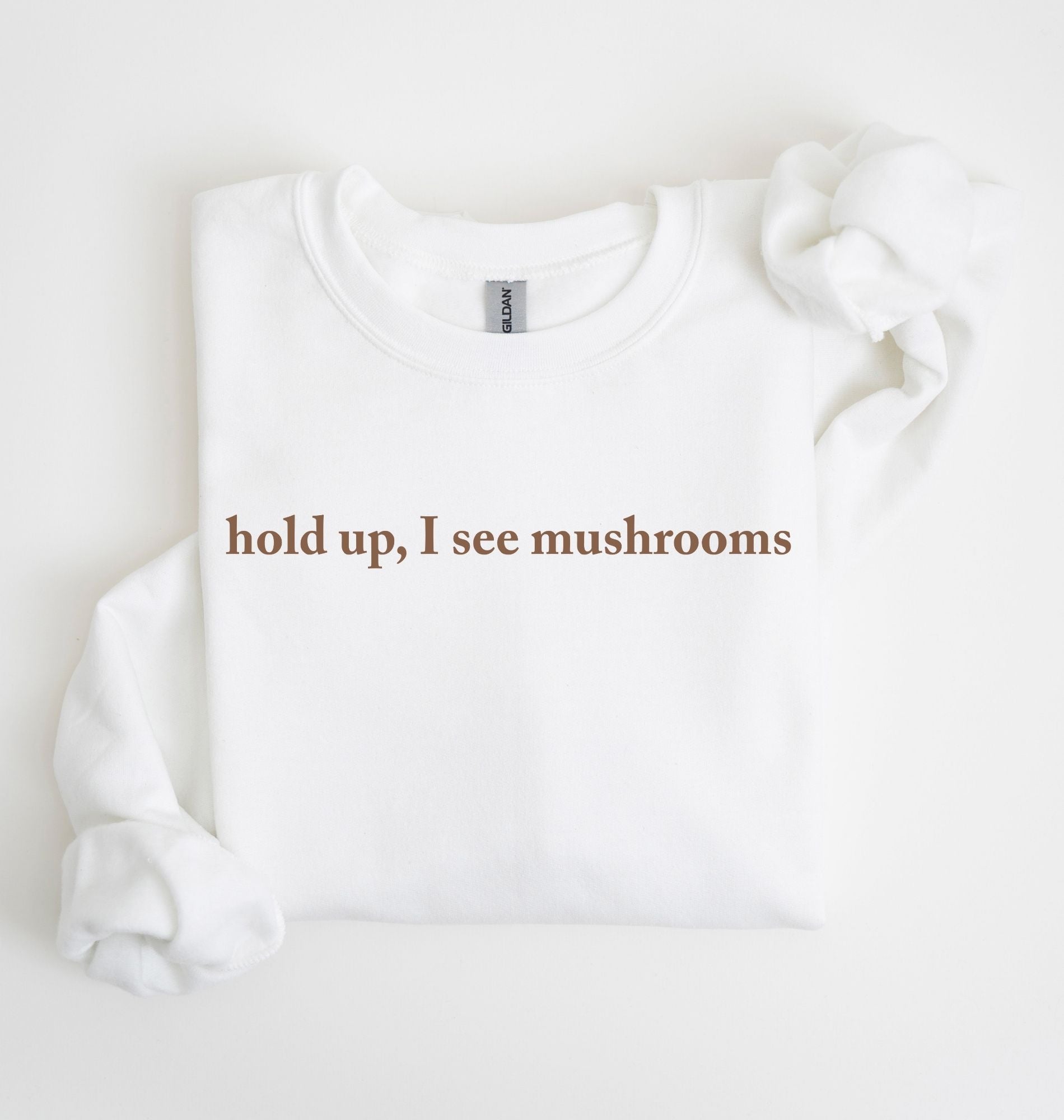 Flat lay of a white-colored sweatshirt on a white background featuring brown text that reads “hold up, I see mushrooms. 