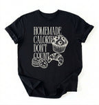 Flat lay of a black Homemade Calories Don't Count baking shirt with muffin cookie and croissant illustrations on a white background 
