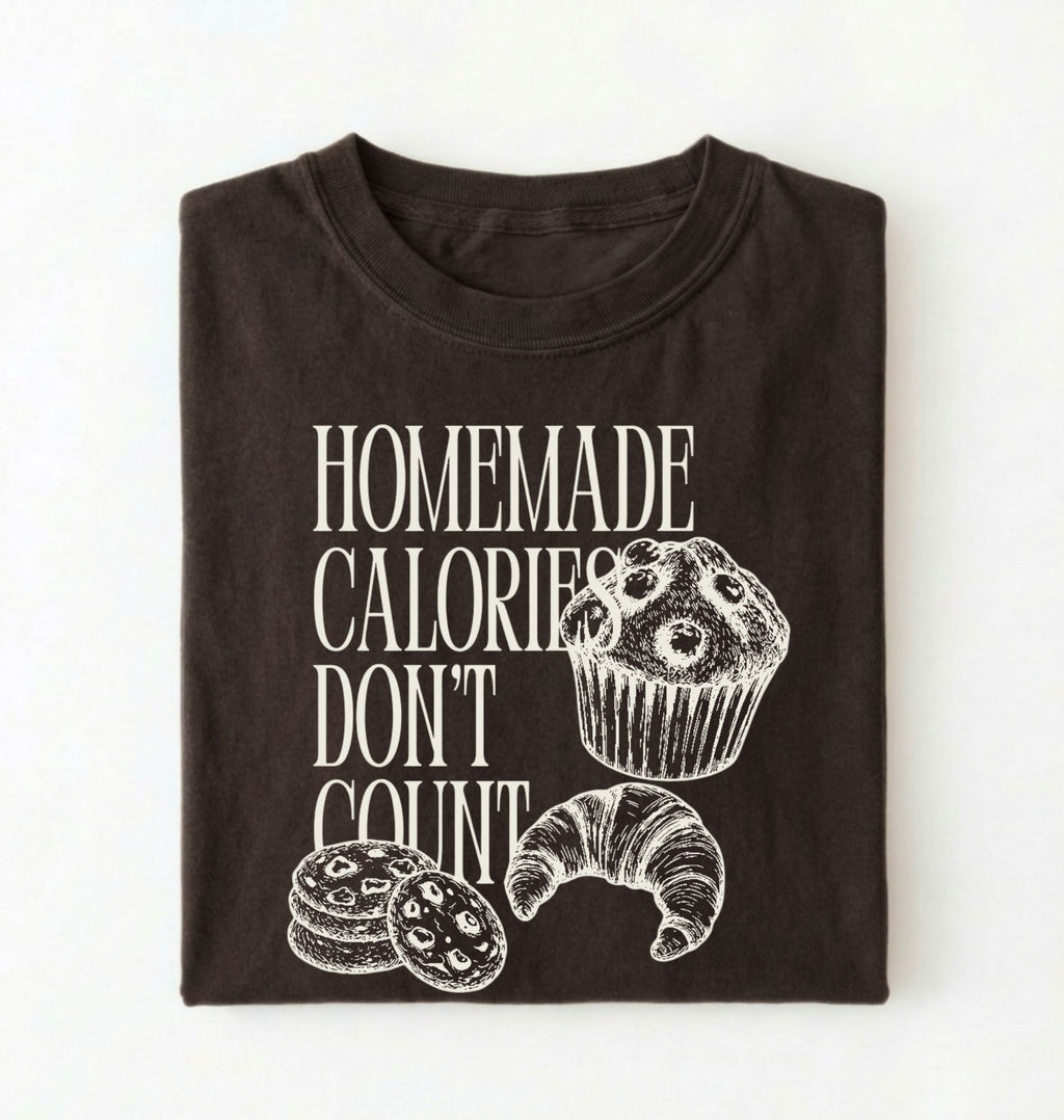 Flat lay of a brown Homemade Calories Don't Count baking shirt with muffin cookie and croissant illustrations on a white background 