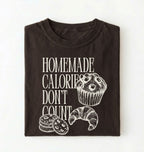 Flat lay of a brown Homemade Calories Don't Count baking shirt with muffin cookie and croissant illustrations on a white background 