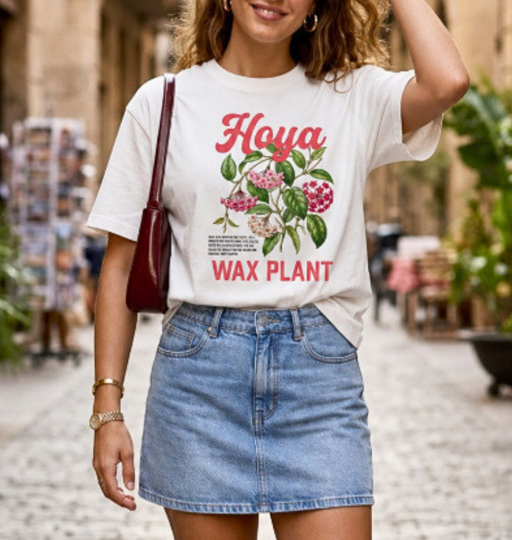 A white graphic t-shirt with the text 'I PLAY IN THE DIRT' and a colorful painting print, styled on a wooden hanger with light blue denim shorts and a brown belt against a white wall.