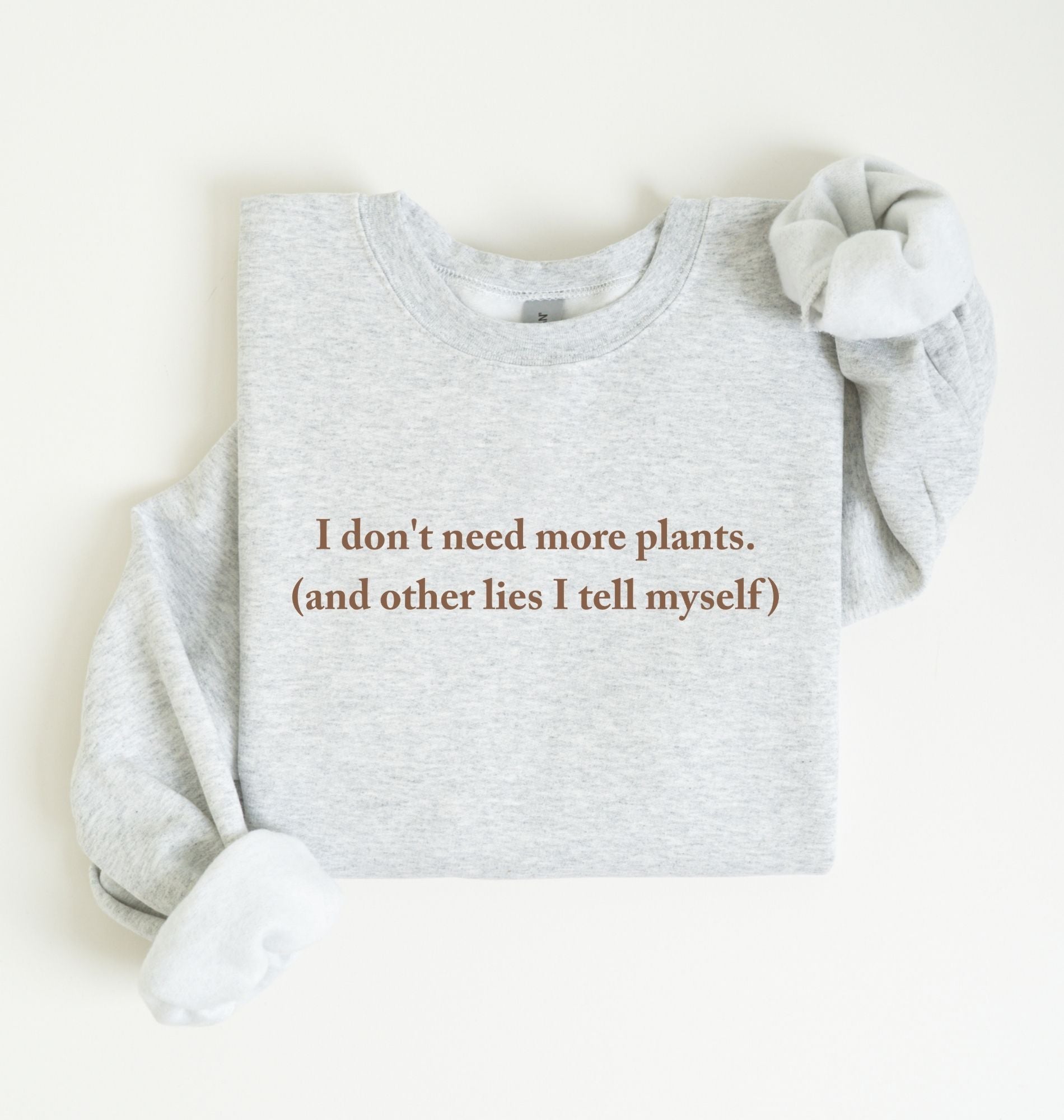 Flat lay of a ash cozy crewneck sweatshirt on a white background with a brown text displaying the phrase “I don’t need more plants (and other lies I tell myself)” centered on the front 