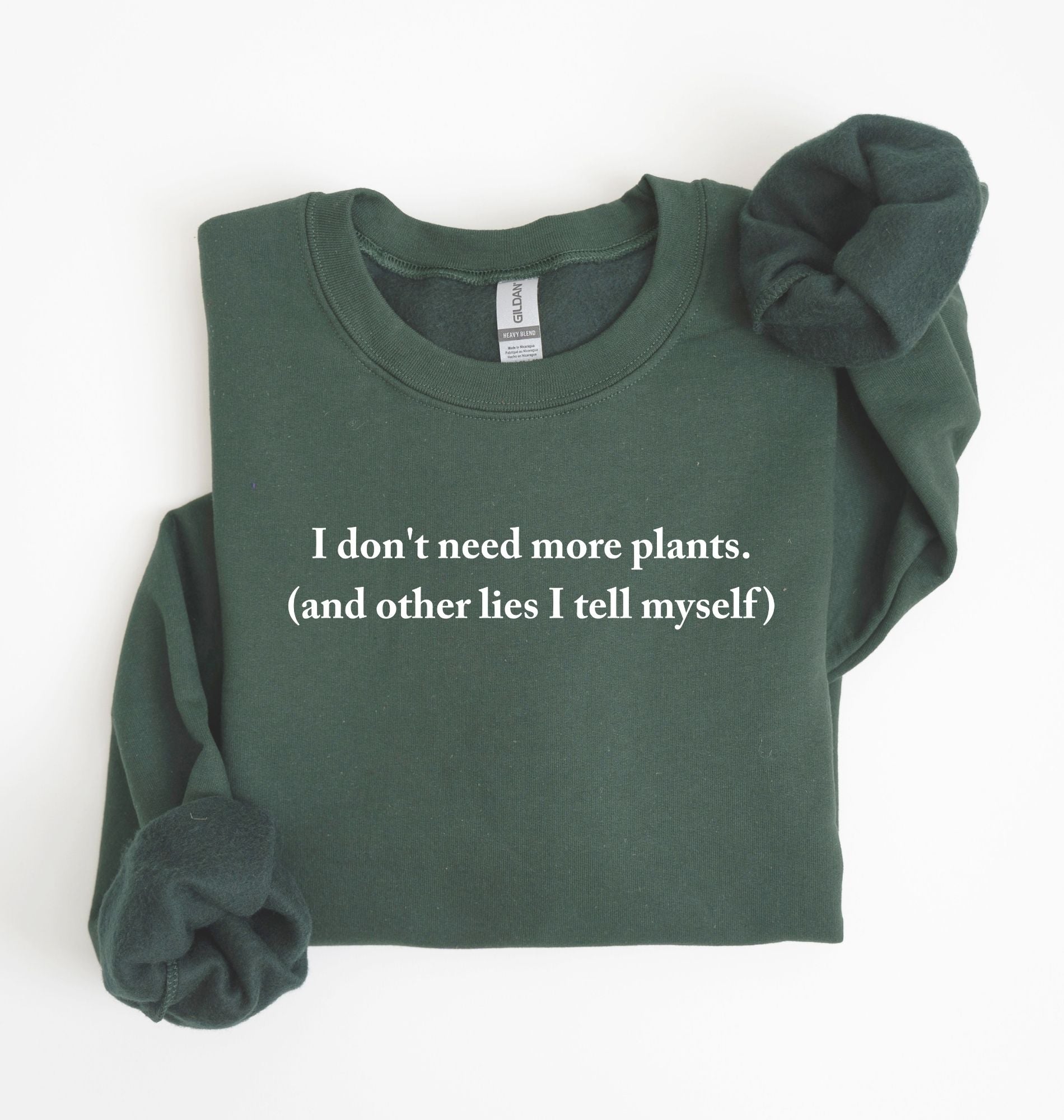Flat lay of a forestgreen cozy crewneck sweatshirt on a white background with a white text displaying the phrase “I don’t need more plants (and other lies I tell myself)” centered on the front 