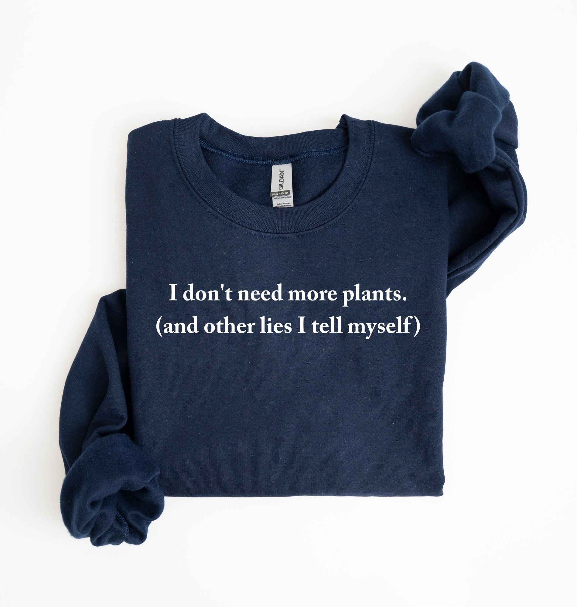 Flat lay of a navyblue cozy crewneck sweatshirt on a white background with a white text displaying the phrase “I don’t need more plants (and other lies I tell myself)” centered on the front 