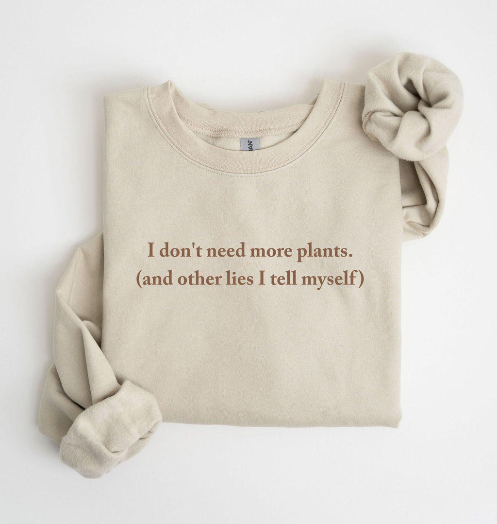 Flat lay of a sand cozy crewneck sweatshirt on a white background with a white text displaying the phrase “I don’t need more plants (and other lies I tell myself)” centered on the front 