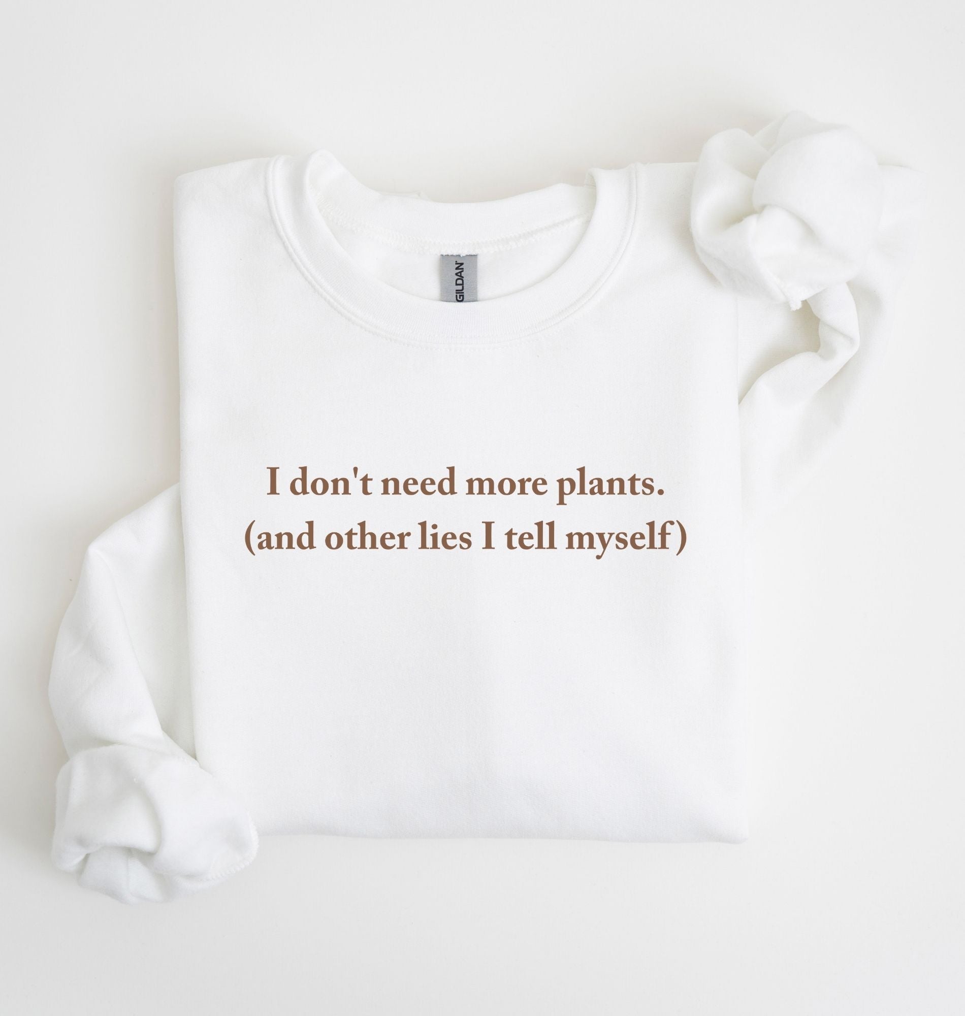 Flat lay of a white cozy crewneck sweatshirt on a white background with a brown text displaying the phrase “I don’t need more plants (and other lies I tell myself)” centered on the front 