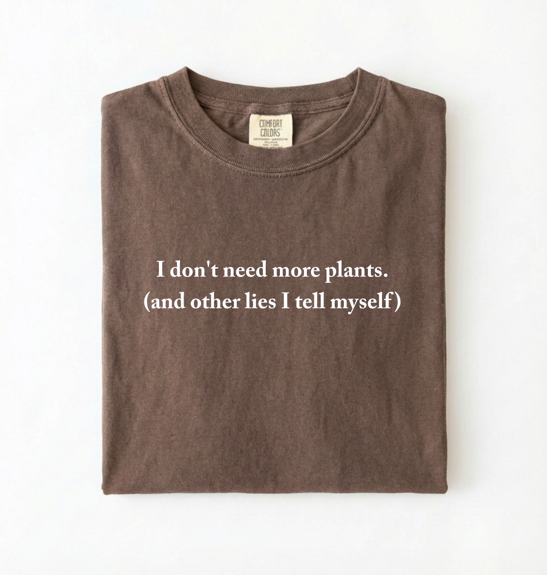 Flat lay of a espresso cozy crewneck t-shirt on a white background with a white text displaying the phrase “I don’t need more plants (and other lies I tell myself)” centered on the front