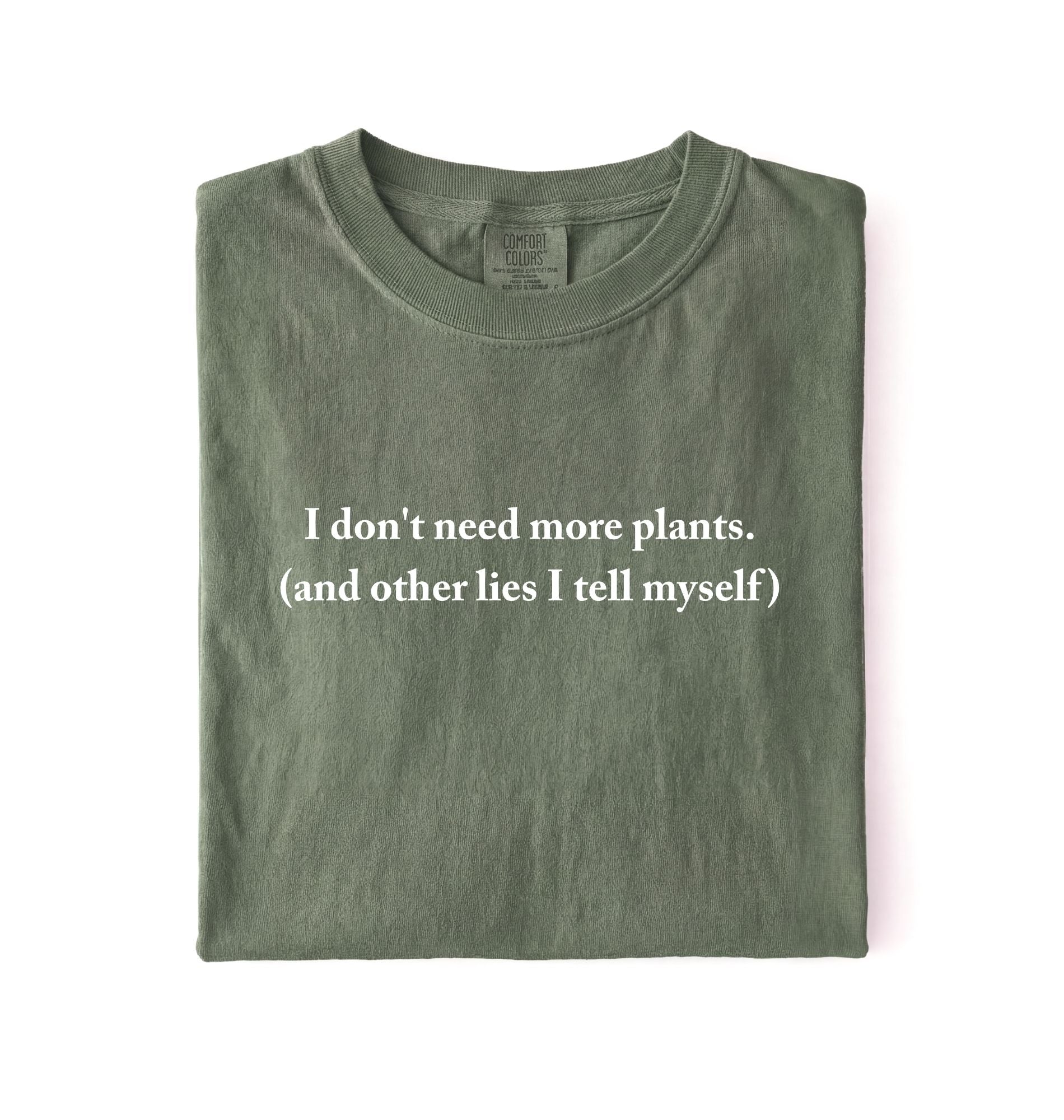 Flat lay of a moss cozy crewneck t-shirt on a white background with a white text displaying the phrase “I don’t need more plants (and other lies I tell myself)” centered on the front