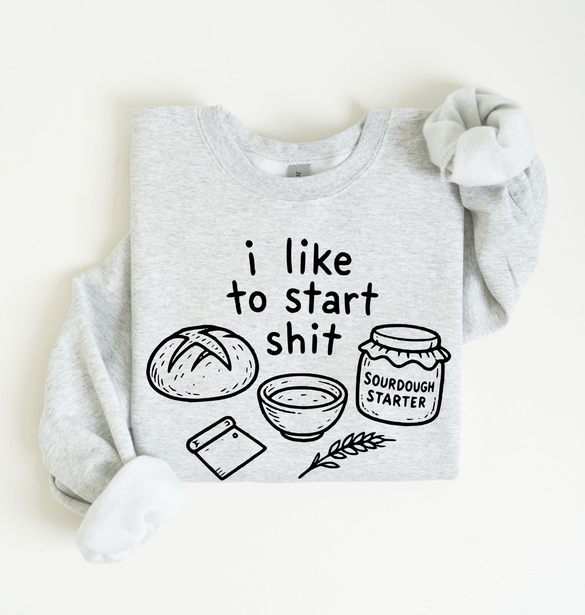 A light heather gray long-sleeve sweatshirt laid flat on a cream background, featuring a black graphic print with the text 'i like to start shit' and illustrations of a loaf of bread, a bowl, a scraper, and a jar labeled 'SOURDOUGH STARTER'.
