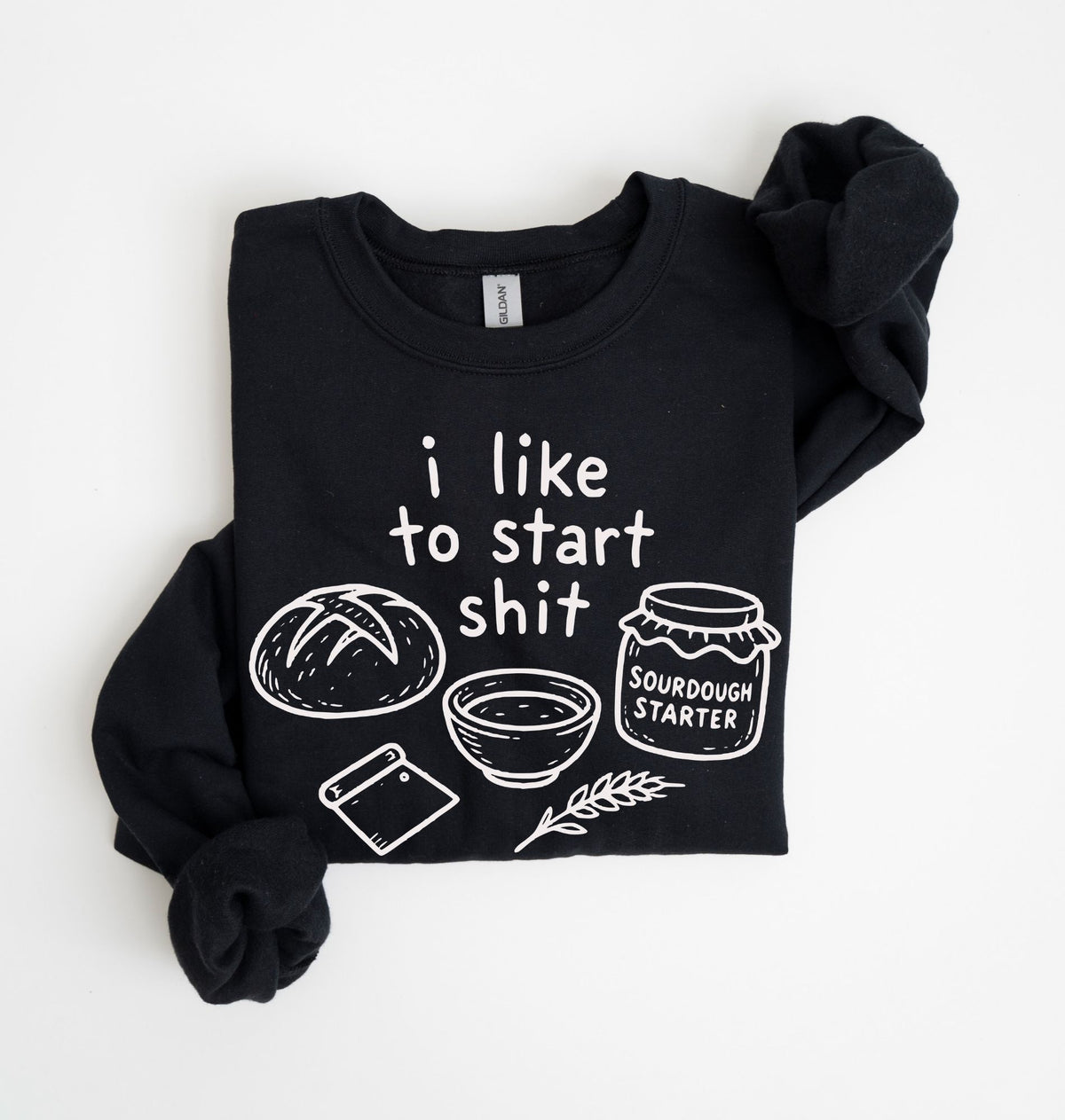 A black long-sleeve sweatshirt with a white graphic print that reads 'i like to start shit' accompanied by drawings of a loaf of bread, a bowl, a bagel, a sprig of dough, and a jar labeled 'SOURDOUGH STARTER'. The sweatshirt is laid flat on a white background with one sleeve rolled up.