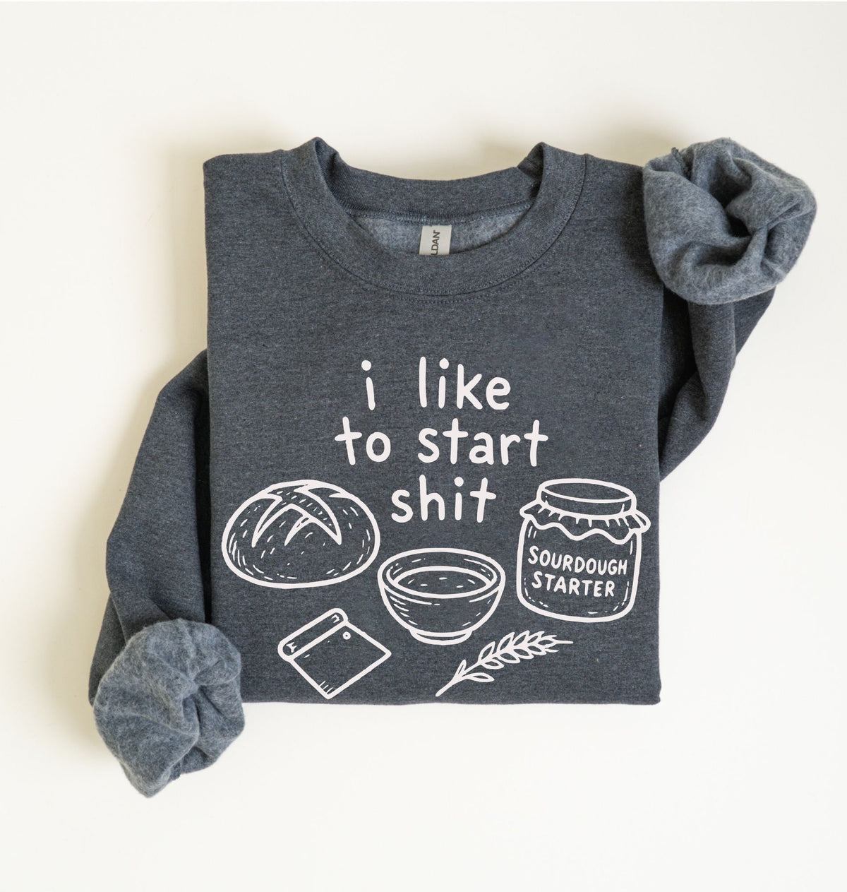 A dark heather gray long-sleeve crewneck sweatshirt with a white graphic print that reads 'i like to start shit' accompanied by drawings of a sourdough bread loaf, a bowl, a knife, a sprig of wheat, and a jar labeled 'SOURDOUGH STARTER'. The sweatshirt is styled with rolled cuffs and a folded sleeve, laid flat on a light cream background.
