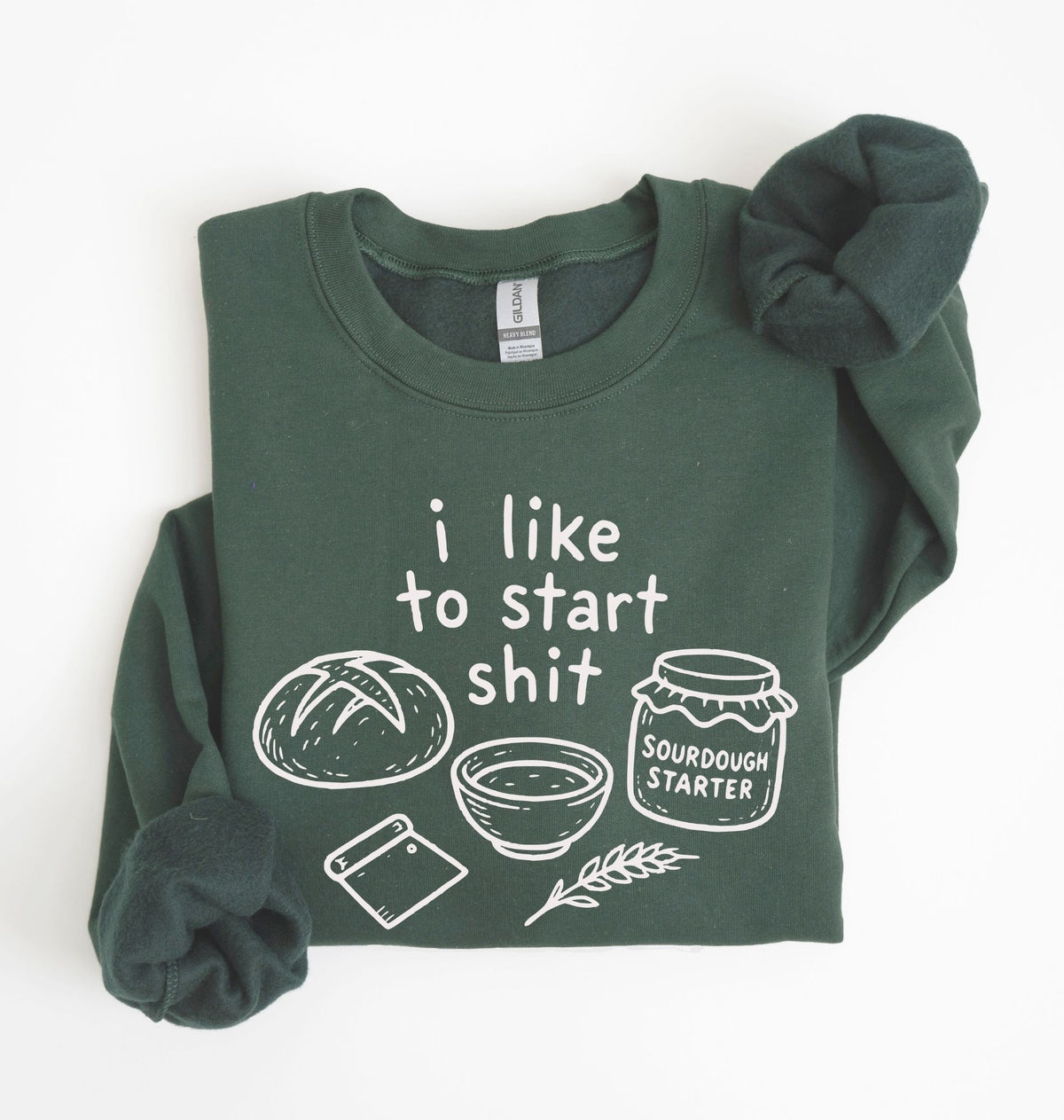 A dark green crewneck sweatshirt with a white graphic print that reads 'i like to start shit' accompanied by illustrations of a sourdough bread loaf, a bowl, a cutting board, and a jar labeled 'SOURDOUGH STARTER'. The sweatshirt is neatly folded on a clean white background with a matching dark green scrunchie.
