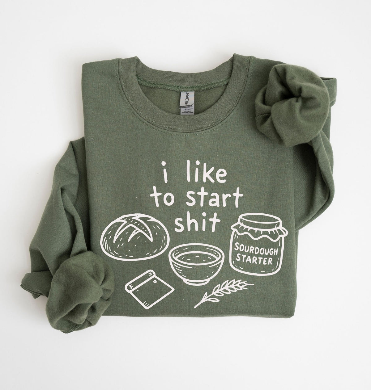 An olive green long-sleeve sweatshirt with a white graphic print that reads 'i like to start shit' accompanied by drawings of a loaf of bread, a bowl, a spatula, and a jar labeled 'SOURDOUGH STARTER'. The sweatshirt is neatly folded with rolled cuffs, set against a clean white background.