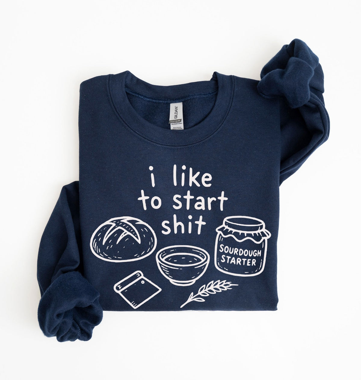 A navy blue long-sleeve t-shirt with a white graphic print that reads 'i like to start shit' accompanied by drawings of a sourdough bread loaf, a bowl, a jar labeled 'SOURDOUGH STARTER', a phone, and a wheat stalk. The shirt is neatly folded on a clean white background.