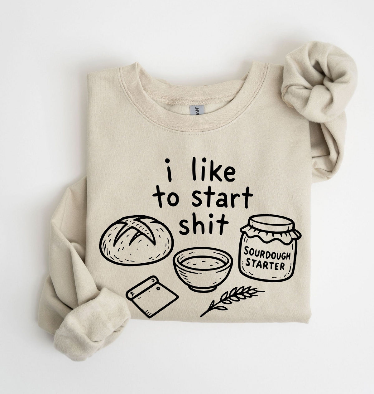 A beige crewneck sweatshirt with a black graphic print that reads 'i like to start shit' above illustrations of a loaf of bread, a bowl, a dough scraper, and a jar labeled 'SOURDOUGH STARTER'. The sweatshirt is styled with rolled sleeves and a matching fabric scrunchie on a clean white background.