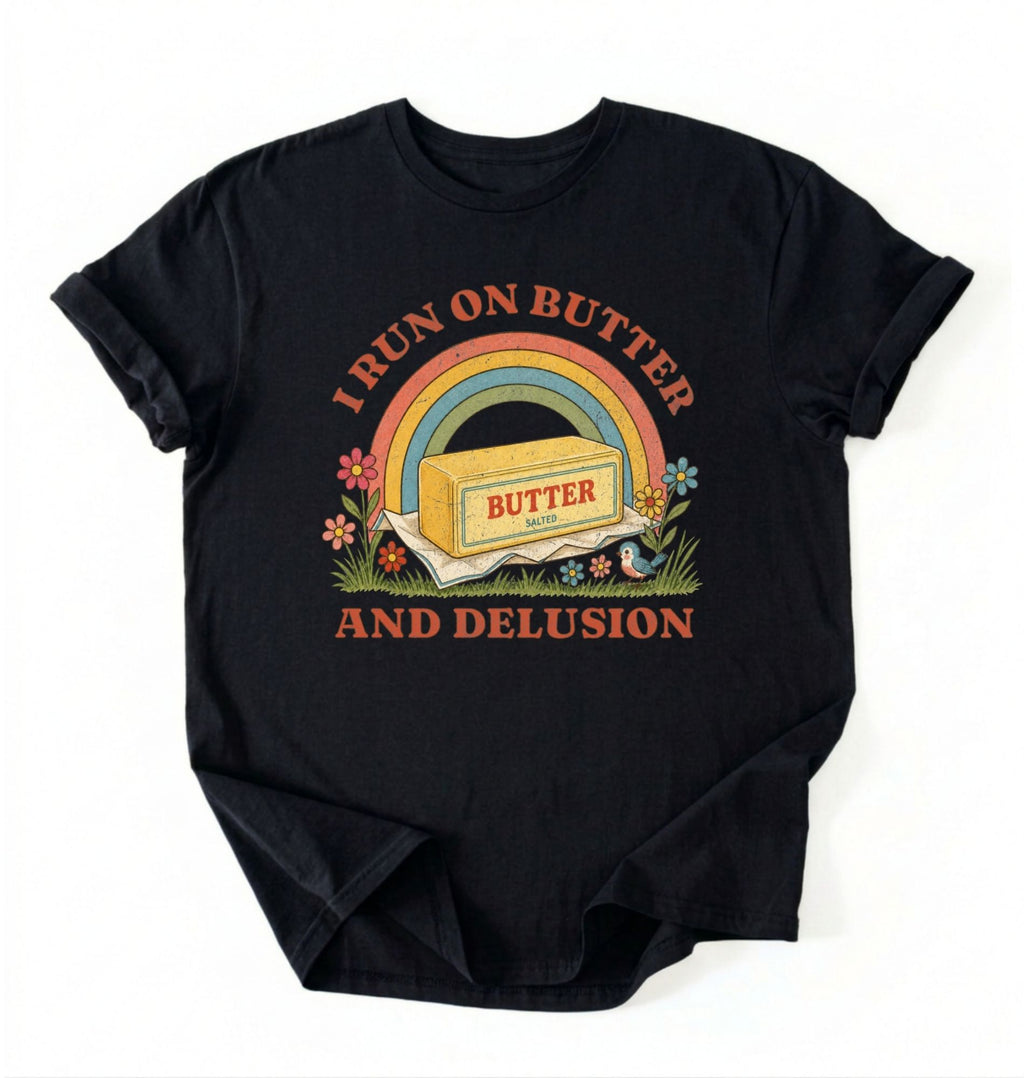 A black short-sleeve t-shirt with a retro-style graphic on the front. The graphic features a rainbow arching over a stick of butter labeled 'BUTTER', with the text 'I RUN ON BUTTER' above and 'AND DELUSION' below in a reddish-brown font, surrounded by small flowers and grass.