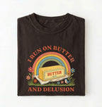 A folded black t-shirt with a retro-style graphic print featuring a yellow butter block under a rainbow, with the text 'I RUN ON BUTTER AND DELUSION' in orange on a white background