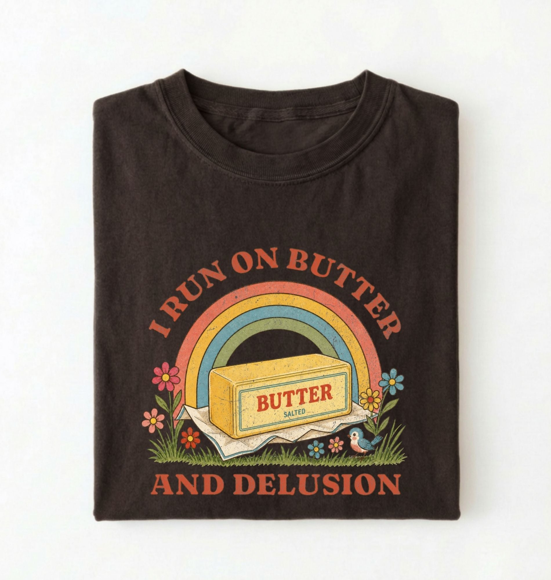 A folded black t-shirt with a retro-style graphic print featuring a yellow butter block under a rainbow, with the text 'I RUN ON BUTTER AND DELUSION' in orange on a white background
