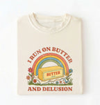 An off-white crewneck t-shirt featuring a retro-style graphic of a butter block under a rainbow with the text 'I RUN ON BUTTER AND DELUSION' in red on a white background