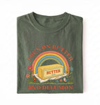 A green short-sleeve t-shirt with a retro-style graphic on the front. The graphic features a rainbow arching over a stick of butter labeled 'BUTTER', with the text 'I RUN ON BUTTER' above and 'AND DELUSION' below in a reddish-brown font, surrounded by small flowers and grass.
