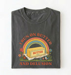 A dark gray folded t-shirt with a retro-style graphic that reads 'I RUN ON BUTTER AND DELUSION' surrounding an illustration of a butter block under a rainbow.