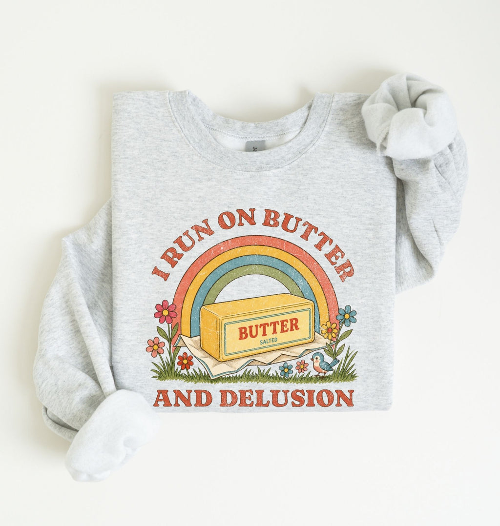 A light heather gray sweatshirt with a retro-style graphic that reads 'I RUN ON BUTTER AND DELUSION' around a rainbow and butter block illustration. The sweatshirt is laid flat on a light cream background with one sleeve rolled up.
