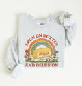 A light heather gray sweatshirt with a retro-style graphic that reads 'I RUN ON BUTTER AND DELUSION' around a rainbow and butter block illustration. The sweatshirt is laid flat on a light cream background with one sleeve rolled up.
