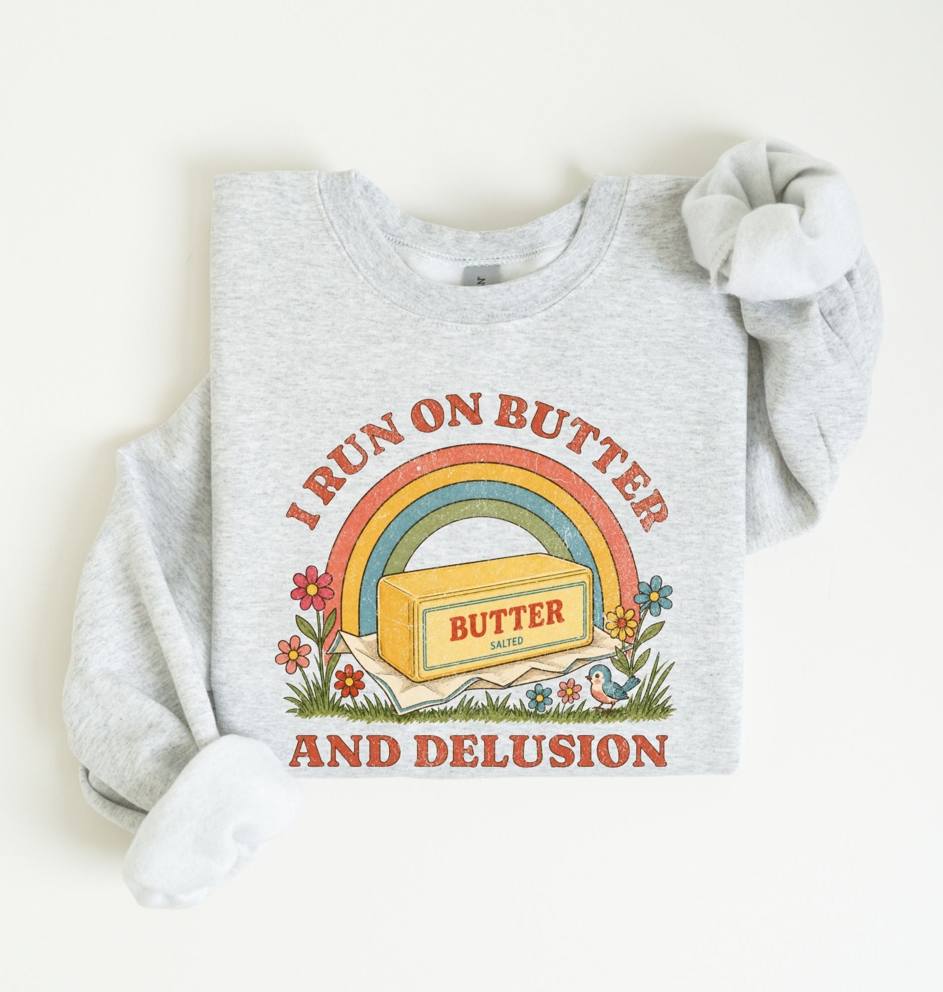 A light heather gray sweatshirt with a retro-style graphic that reads 'I RUN ON BUTTER AND DELUSION' around a rainbow and butter block illustration. The sweatshirt is laid flat on a light cream background with one sleeve rolled up.