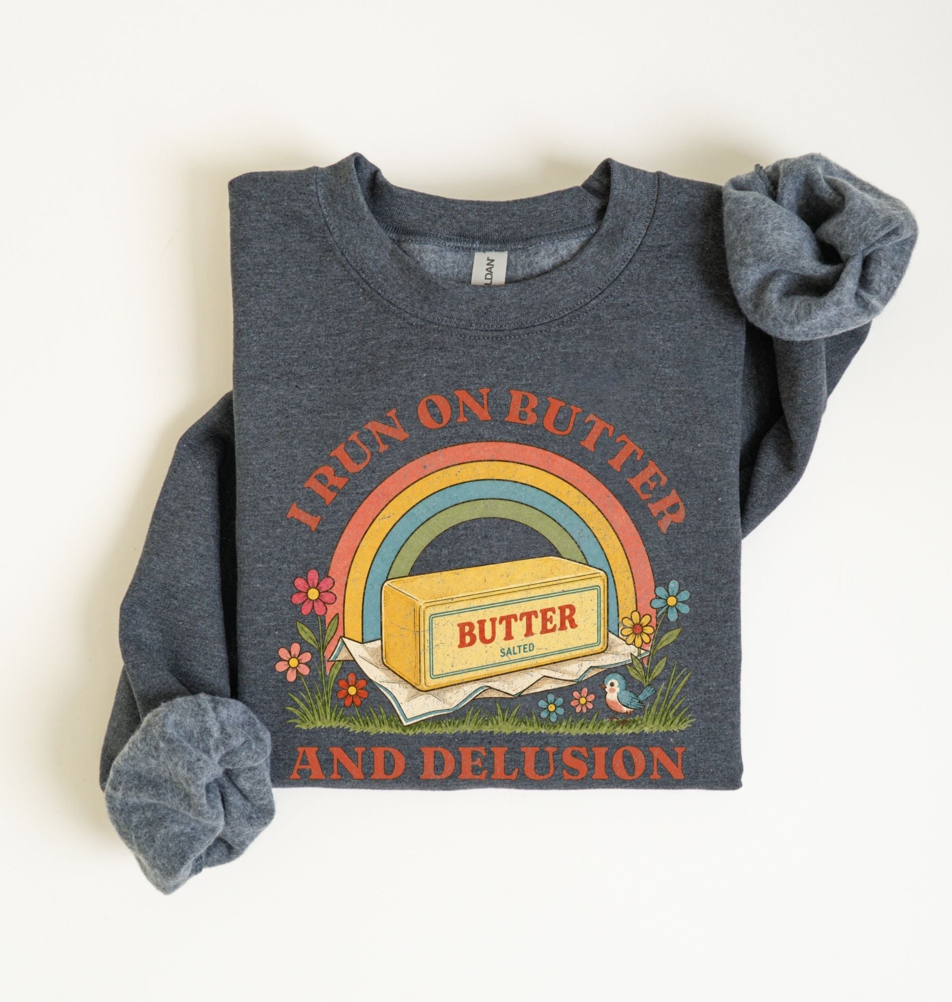 A dark heather gray long-sleeve t-shirt with rolled cuffs, featuring a retro graphic of a butter block under a rainbow with the text 'I RUN ON BUTTER AND DELUSION' on a white background