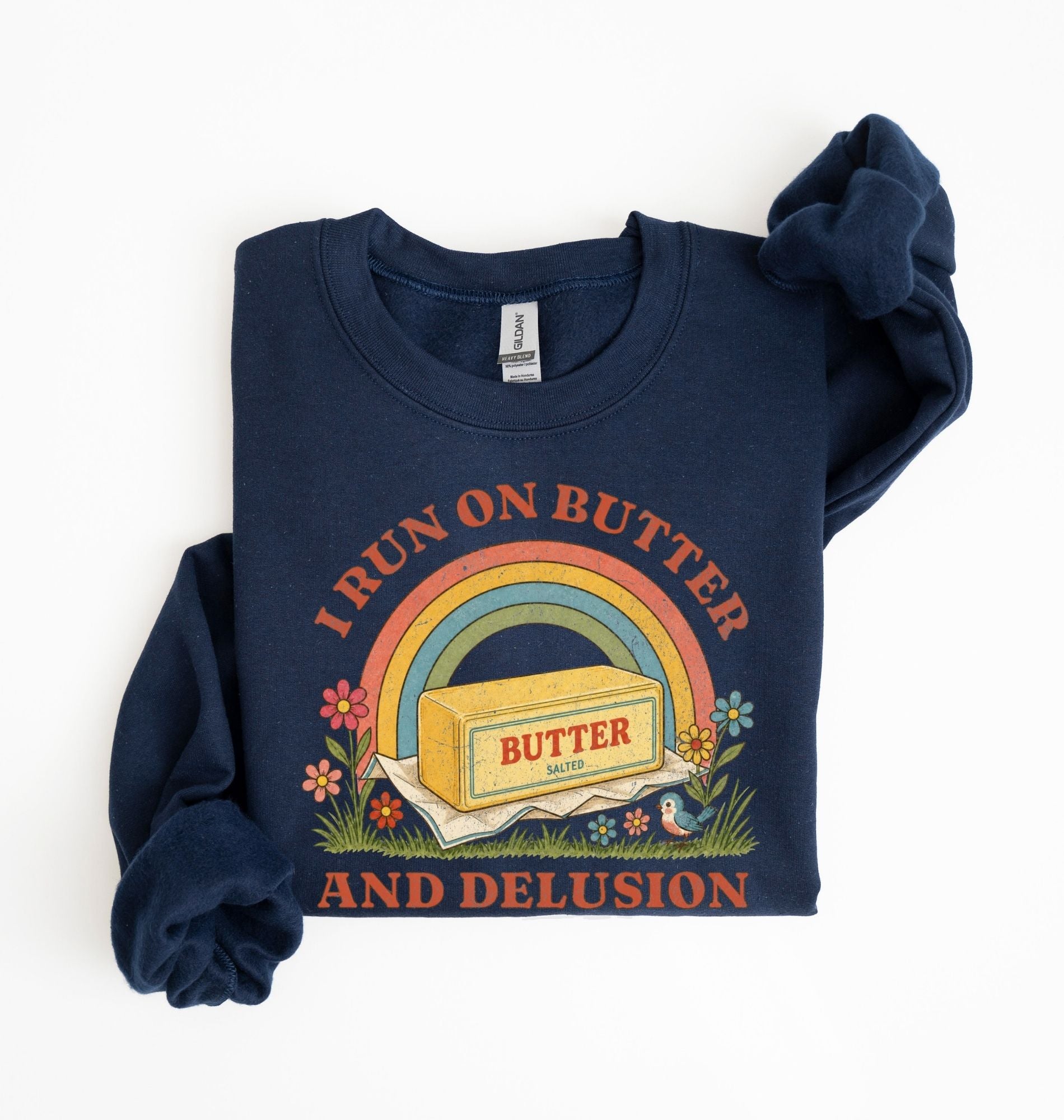 A navy blue long-sleeve t-shirt with a retro-style graphic print. The graphic features a yellow stick of butter with 'BUTTER' written on it, placed under a rainbow and surrounded by flowers, with the text 'I RUN ON BUTTER AND DELUSION' in orange arched above and below.