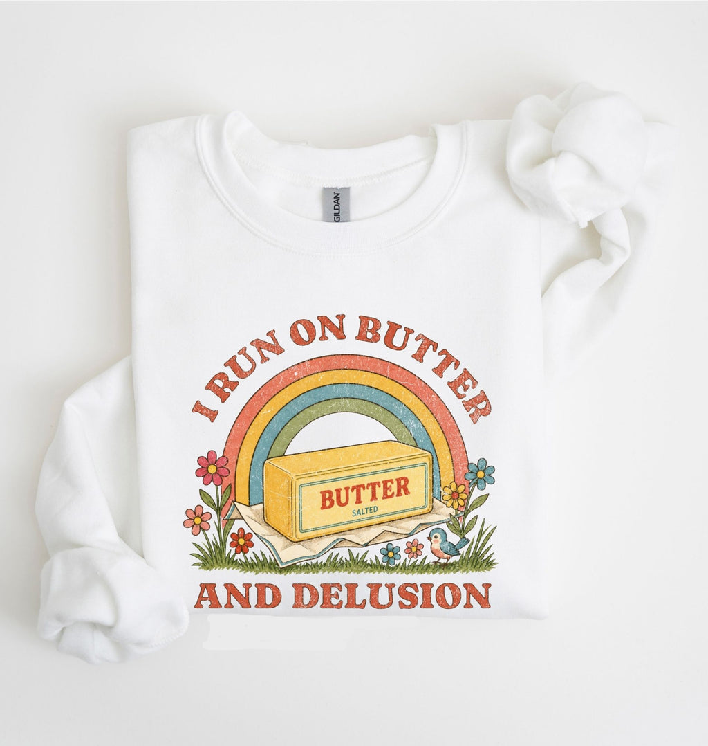 A white long-sleeve sweatshirt with a retro-style graphic print on the front. The graphic features a yellow butter block under a rainbow with the text 'I RUN ON BUTTER AND DELUSION' in a reddish-brown font, surrounded by small colorful flowers.