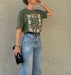 A woman models an olive green graphic t-shirt featuring a vintage-style illustration of kitchen utensils and the text 'i'll be in my office'. She is wearing light-wash blue jeans, a black belt, and holding a small black clutch against a textured gray wall.