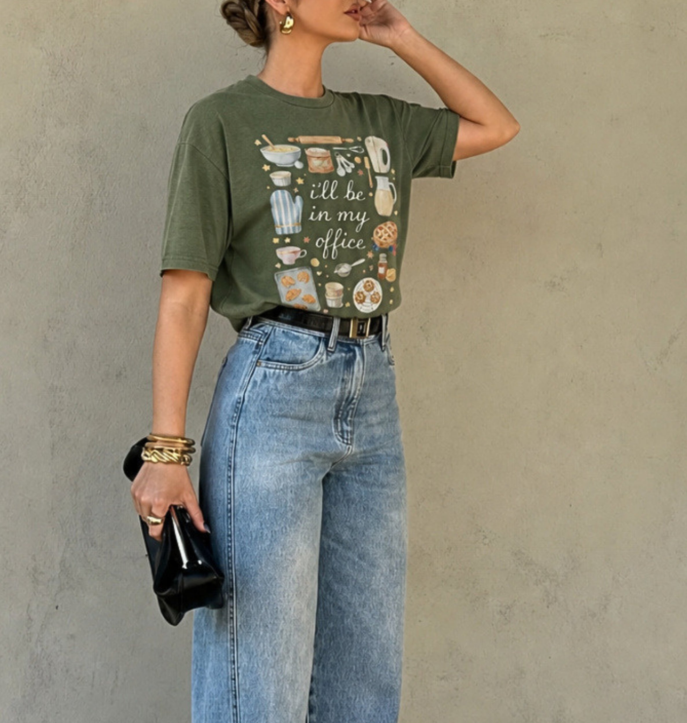 A woman models an olive green graphic t-shirt featuring a vintage-style illustration of kitchen utensils and the text 'i'll be in my office'. She is wearing light-wash blue jeans, a black belt, and holding a small black clutch against a textured gray wall.