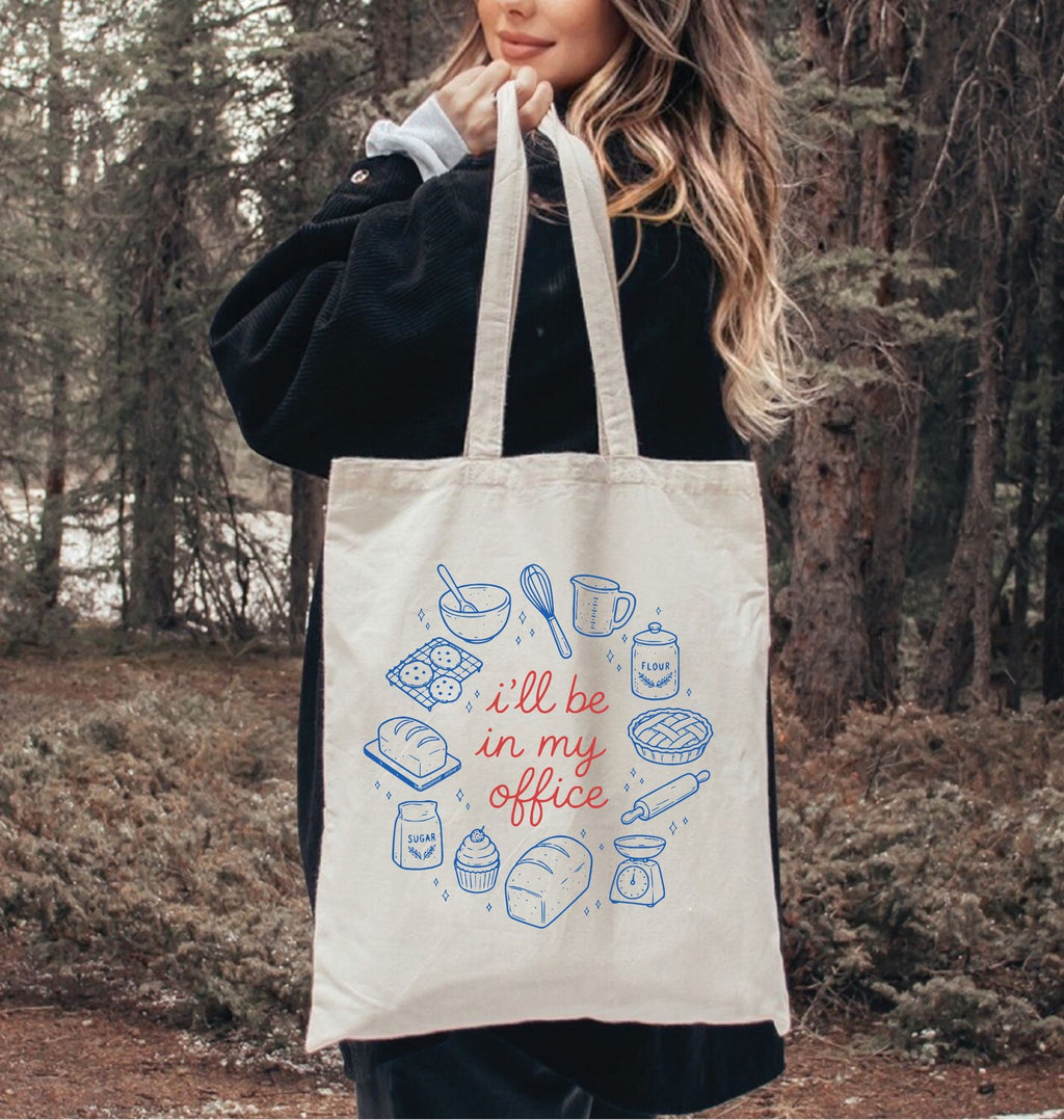 Person holding a tote bag with baking illustrations and text in a forest setting