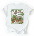 Flat lay of a white t-shirt featuring illustrated potted plants, tools, and text reading “I’ll Be In My Office Garden Club on a white background