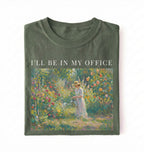 Flat lay of a moss t-shirt featuring a vintage garden painting of a woman tending flowers with text reading “I’ll Be In My Office.” On a white background 