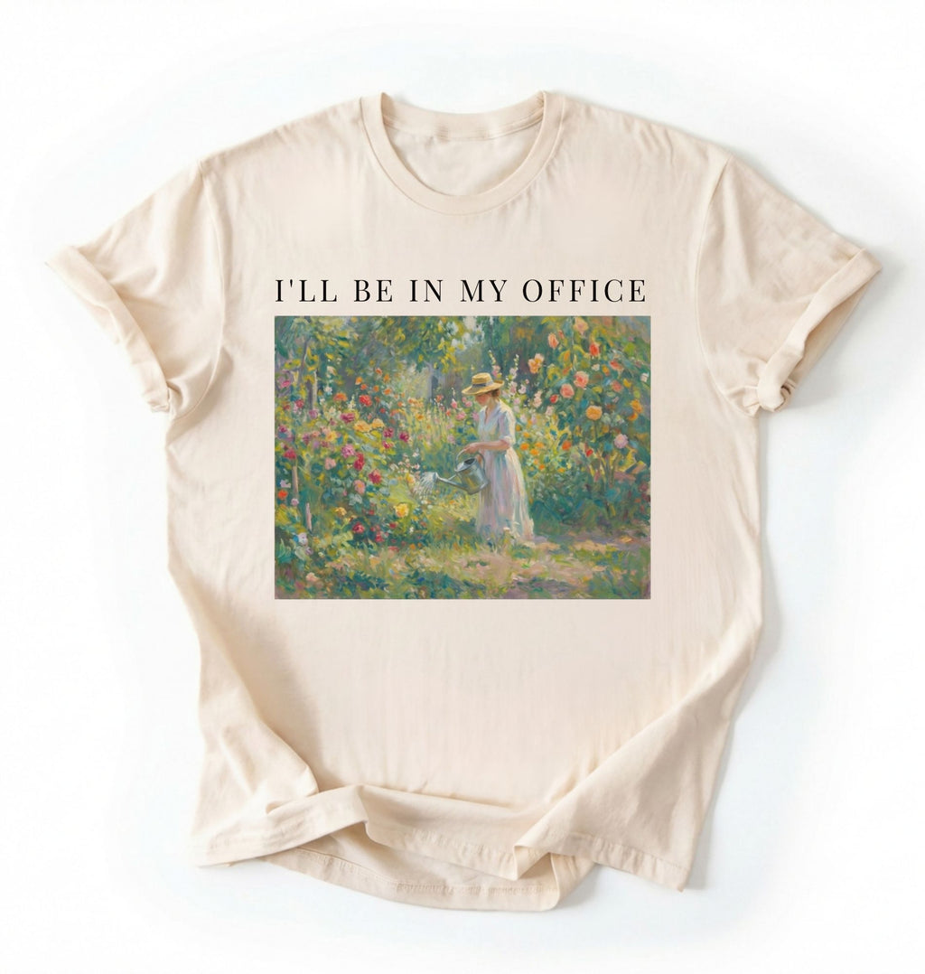 Flat lay of a natural t-shirt featuring a vintage garden painting of a woman tending flowers with text reading “I’ll Be In My Office.” On a white background 