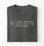 Flat lay of a dark grey t-shirt featuring text reading “I’m a great gardener, that plant didn’t try hard enough.” Funny gardening shirt perfect for plant lovers, gardeners and houseplant owners on a white background