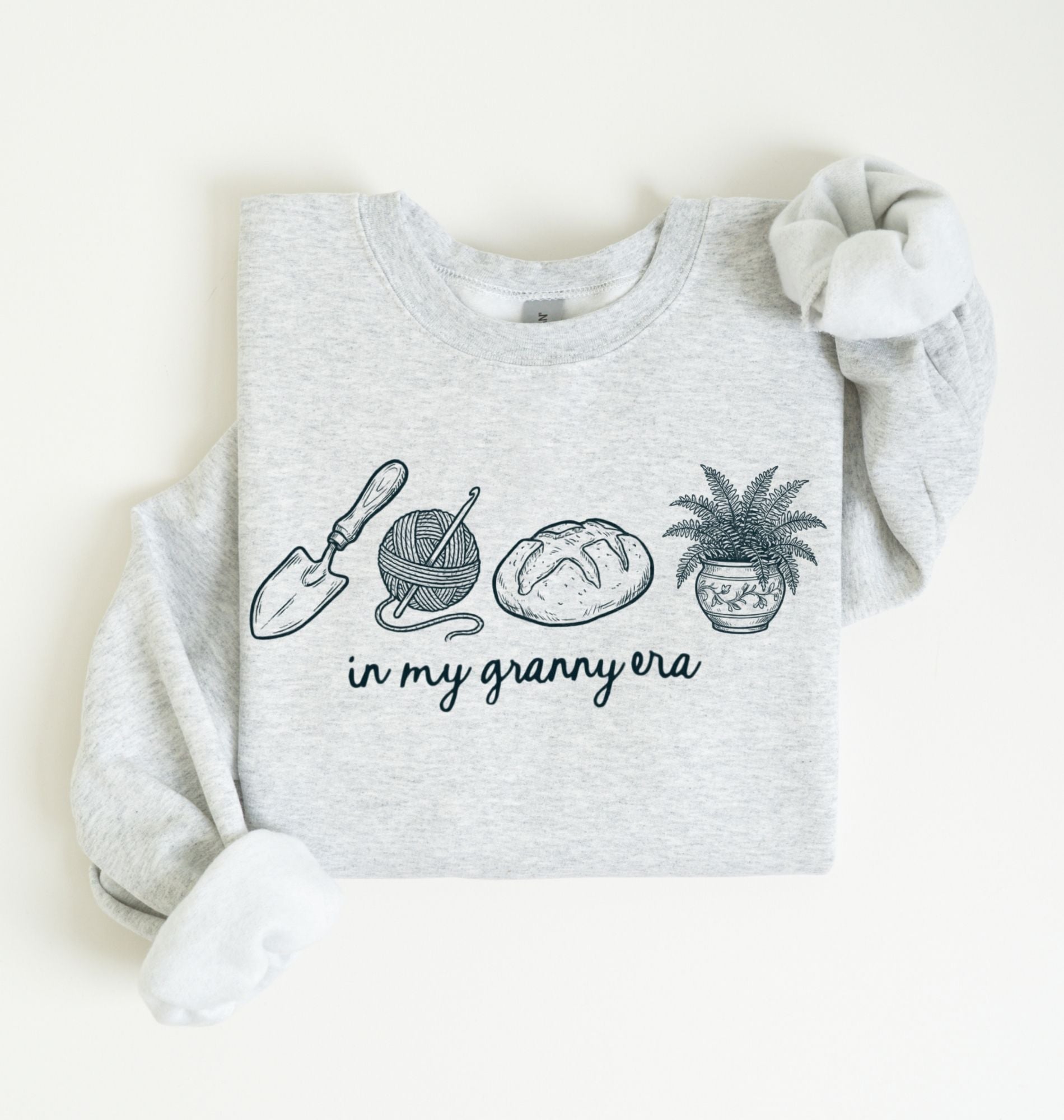 Flat lay of a ash crewneck sweatshirt with line drawings of gardening items and text reading “in my granny era.”
