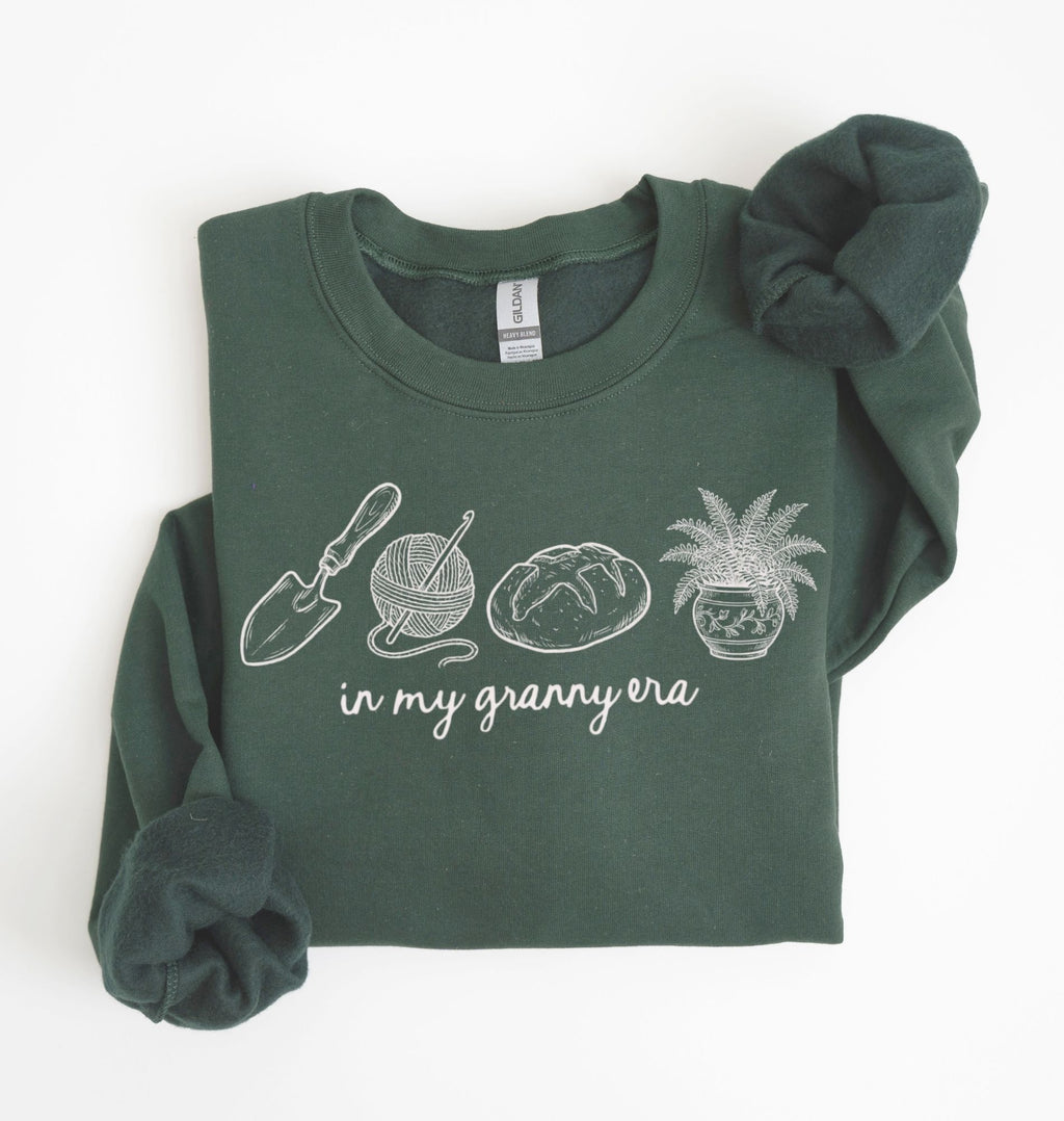 Flat lay of a forestgreen crewneck sweatshirt with line drawings of gardening items and text reading “in my granny era.”