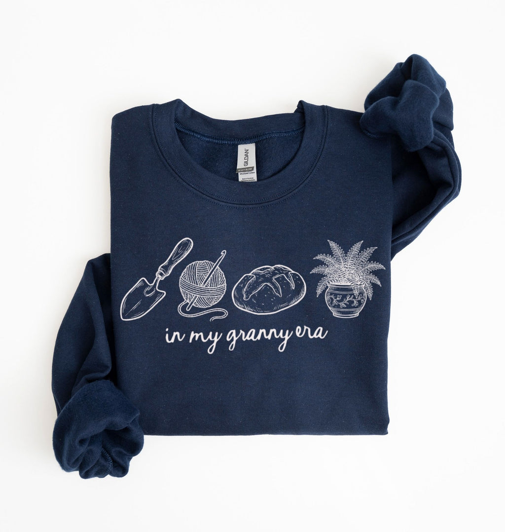 Flat lay of a navy blue crewneck sweatshirt with line drawings of gardening items and text reading “in my granny era.”