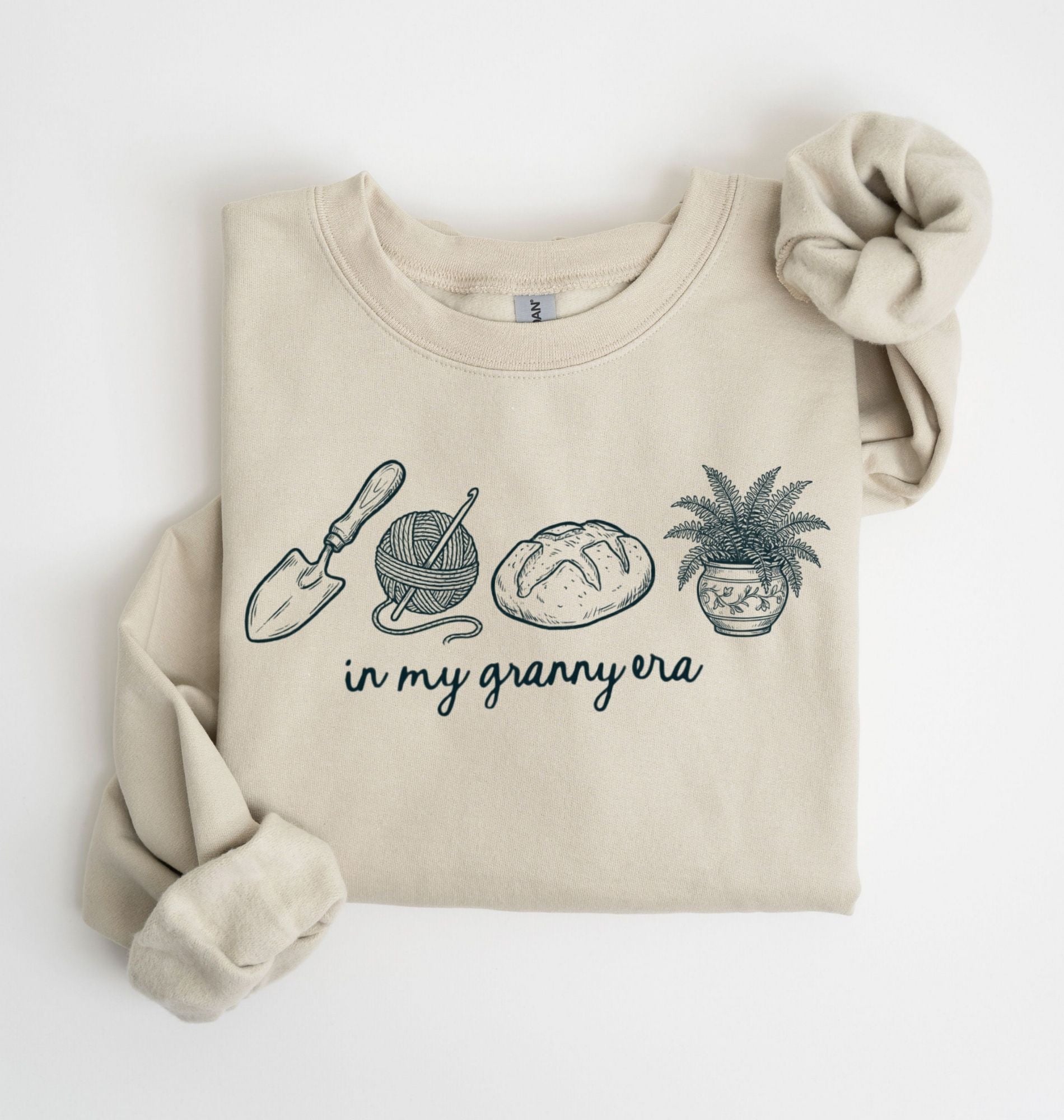 Flat lay of a sand crewneck sweatshirt with line drawings of gardening items and text reading “in my granny era.”
