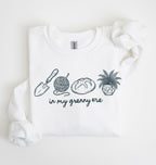 Flat lay of a white crewneck sweatshirt with line drawings of gardening items and text reading “in my granny era.”
