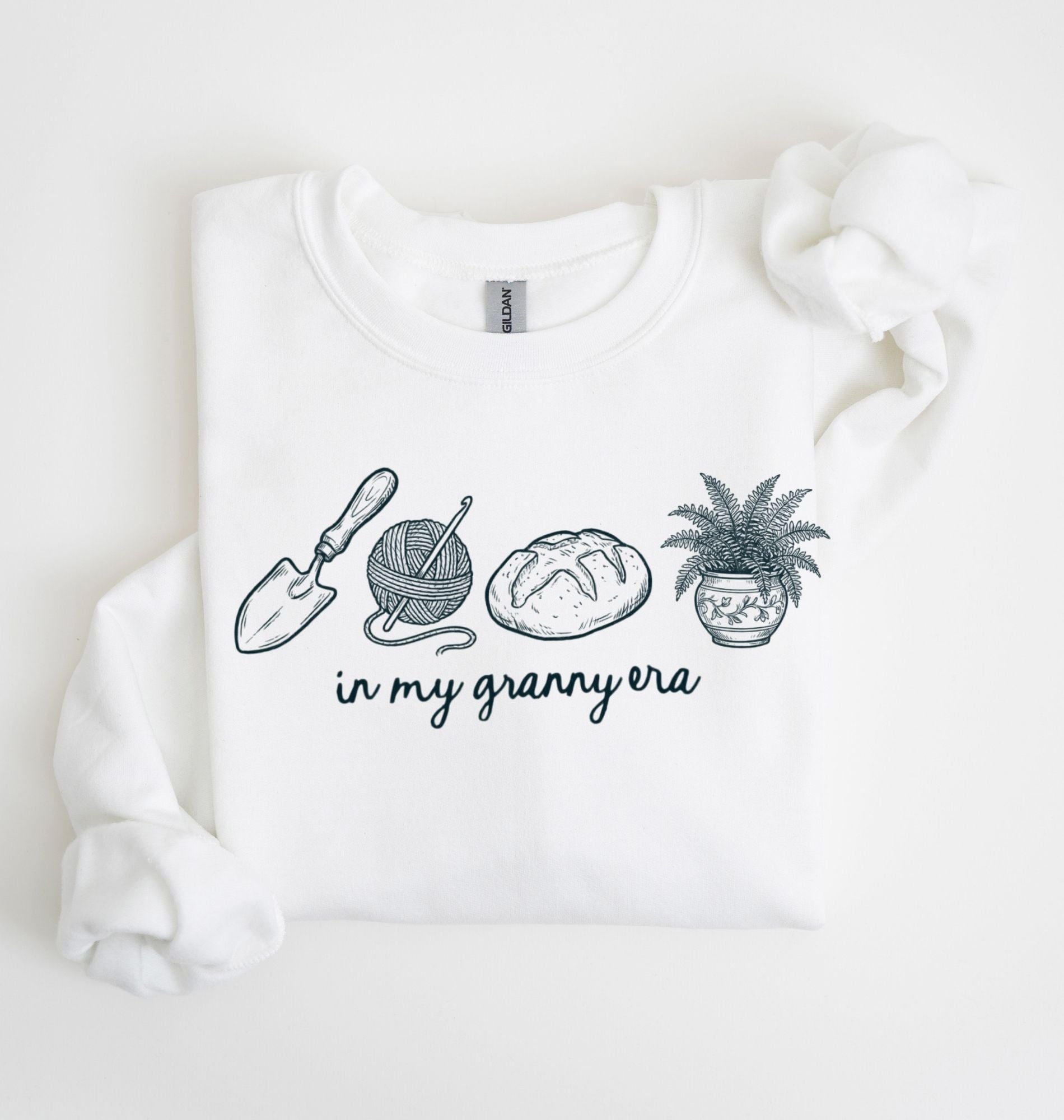Flat lay of a white crewneck sweatshirt with line drawings of gardening items and text reading “in my granny era.”